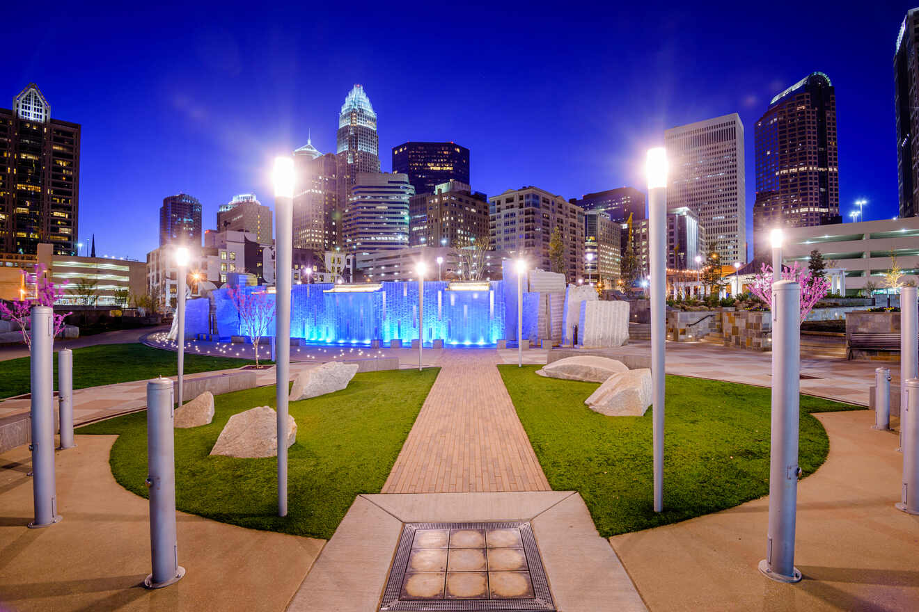 Nighttime view of a park in Charlotte with illuminated water fountains, walking paths, and the city skyline in the background